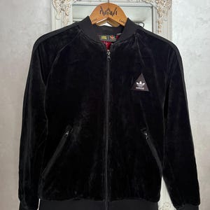 May include: Black velvet bomber jacket with a zip-up front, side pockets, and a ribbed hem and cuffs. Features the Adidas logo on the chest. The jacket is hanging on a wooden hanger.