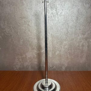 May include: A silver metal stand with a round base and a long, thin rod extending upwards. The rod has a small hook at the top.