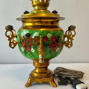 May include: A gold-colored samovar with a green and red floral design. The traditional tea kettle features ornate handles and a spigot. The electrical cord is visible. A decorative piece for the kitchen.