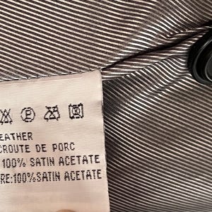 Peut inclure: Gros plan sur l'int&eacute;rieur d'un v&ecirc;tement, avec une &eacute;tiquette indiquant les symboles d'entretien et le texte. L'&eacute;tiquette indique "REAL LEATHER" et d&eacute;taille la composition de la doublure. Un bouton noir est visible sur le tissu ray&eacute;.