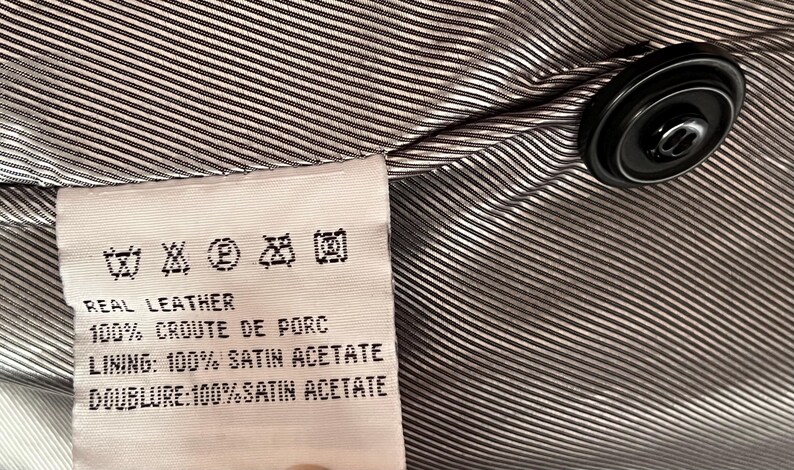 Peut inclure: Gros plan sur l'int&eacute;rieur d'un v&ecirc;tement, avec une &eacute;tiquette indiquant les symboles d'entretien et le texte. L'&eacute;tiquette indique "REAL LEATHER" et d&eacute;taille la composition de la doublure. Un bouton noir est visible sur le tissu ray&eacute;.