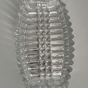 Puede incluir: Un cenicero rectangular de vidrio transparente con un patrón geométrico texturizado. El cenicero tiene una serie de diseños cuadrados en relieve en el interior y exterior, creando una apariencia facetada. Los bordes son ligeramente festoneados.