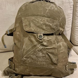 Könnte beinhalten: Ein brauner Rucksack aus Segeltuch mit einer großen, mittigen Außentasche mit Knopfverschluss, verstellbaren Seitengurten mit Metallschnallen und einer Kordelzugöffnung oben. Der Stoff weist eine verwitterte, strukturierte Optik auf.