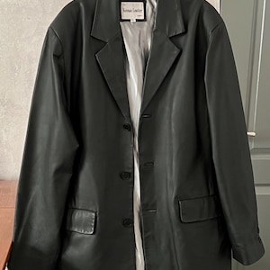Peut inclure: Un blazer en cuir noir de conception classique, avec un revers crant&eacute;, deux poches avant et une fermeture &agrave; trois boutons. Le blazer est suspendu &agrave; un cintre. L'&eacute;tiquette indique "Keenan Leather".