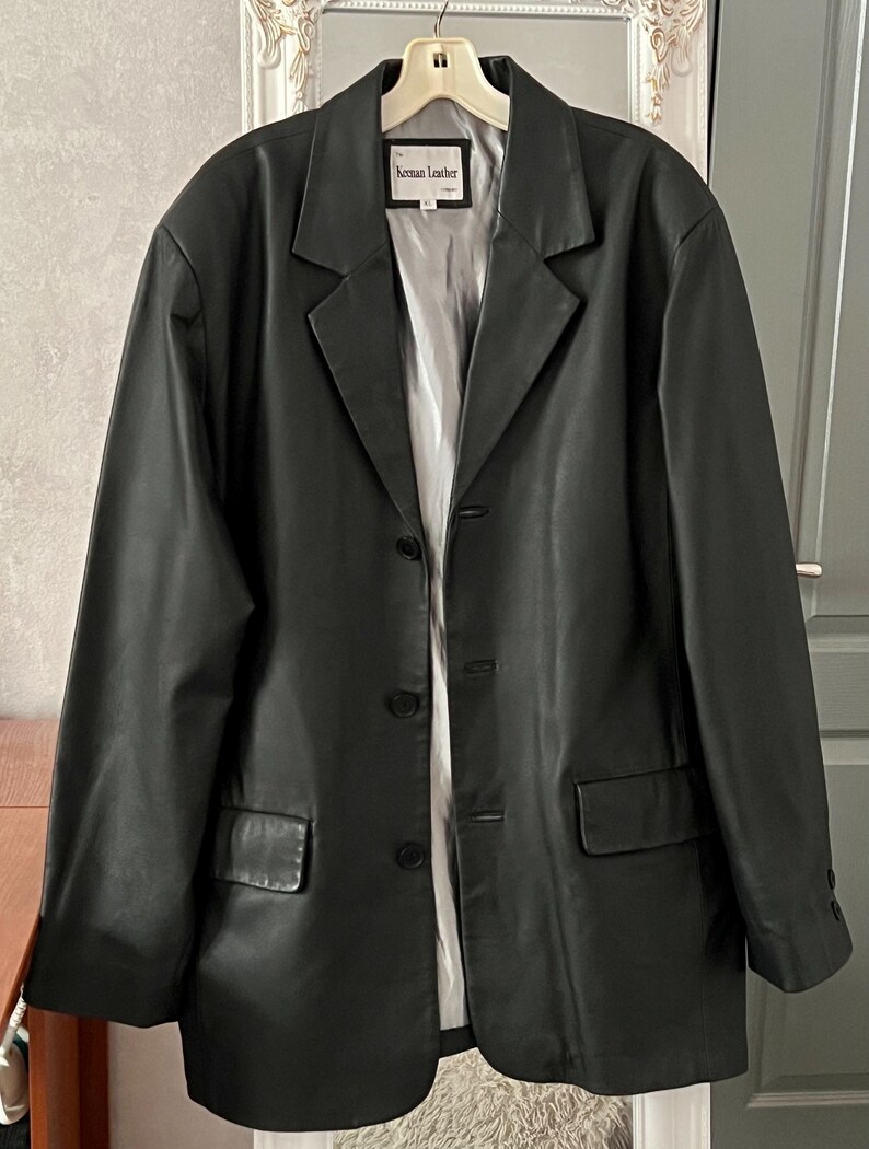 Peut inclure: Un blazer en cuir noir de conception classique, avec un revers crant&eacute;, deux poches avant et une fermeture &agrave; trois boutons. Le blazer est suspendu &agrave; un cintre. L'&eacute;tiquette indique "Keenan Leather".