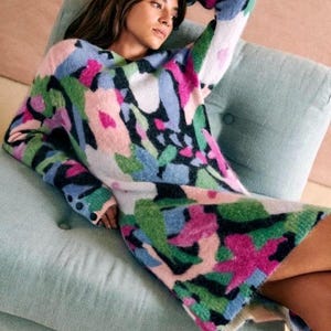 May include: A long-sleeved sweater dress featuring a vibrant floral pattern in pink, green, blue, and white. The dress has a crew neck and is knee-length. The model is seated on a light blue cushioned chair.