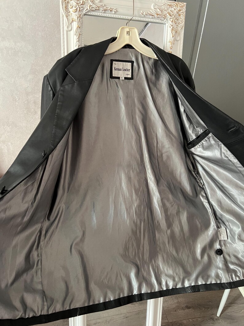 Peut inclure: Veste en cuir noir avec une doublure en satin gris. La veste est ouverte, r&eacute;v&eacute;lant la doublure int&eacute;rieure et une petite &eacute;tiquette qui indique "Keman Leather". La veste est suspendue &agrave; un cintre blanc.