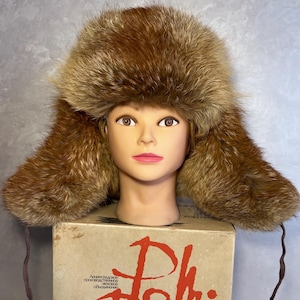 May include: A brown fur hat with ear flaps and a rounded crown. The hat is displayed on a mannequin head. The hat has a vintage look and is likely made of fox fur. The hat is sitting on a cardboard box with text.