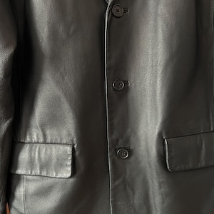 Peut inclure: Gros plan sur un blazer en cuir noir avec trois boutons et deux poches avant. Le blazer a un design classique et une texture lisse. Les boutons sont noirs et ronds.