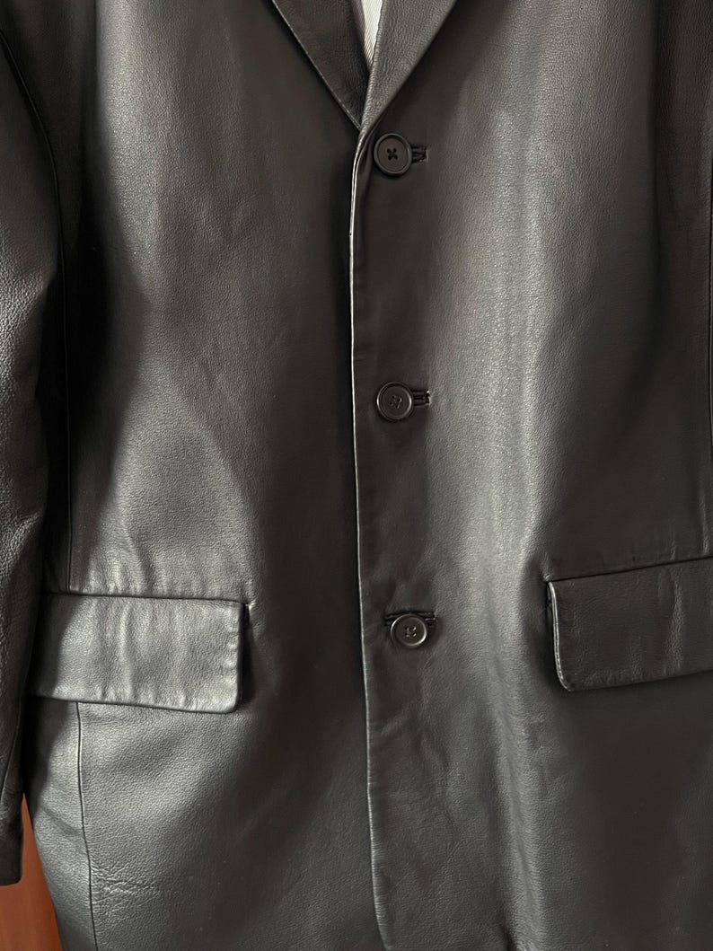 Peut inclure: Gros plan sur un blazer en cuir noir avec trois boutons et deux poches avant. Le blazer a un design classique et une texture lisse. Les boutons sont noirs et ronds.