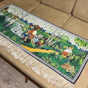 Puede incluir: Un camino de mesa rectangular de tela con una colorida ilustración de gatos antropomórficos en un entorno forestal. La escena incluye gatos pescando, caminando y abedules, bordeado por un patrón negro y acabado con flecos blancos.