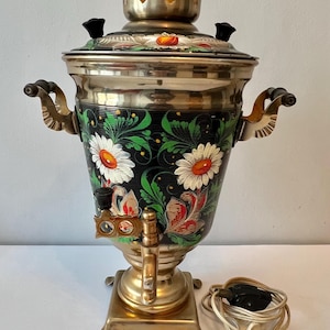 May include: A white ceramic mug with a black handle and the text "But First, Coffee" in a bold, black font. The mug is set against a blurred background, suggesting a cozy setting. The mug appears to be approximately 4 inches tall.