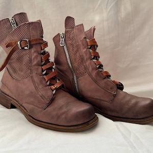 May include: A pair of dusty rose leather boots with a distressed finish. The boots have a side zip, perforated detailing, and brown leather straps with buckles. The boots have brown laces and a low heel.