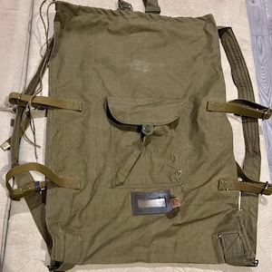 May include: An olive green canvas rucksack featuring a drawstring top closure, adjustable shoulder straps, and side compression straps. A central pocket with a buttoned flap is positioned above a rectangular brown leather identification tag holder.