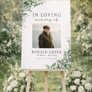 Peut inclure: Un panneau comm&eacute;moratif sur un chevalet en bois, avec le texte "IN LOVING memory of" au-dessus d'une photo d'un homme. Le panneau comprend le nom "RONALD GREEN" et des dates, avec des accents floraux. Des fleurs blanches et de la verdure entourent le panneau.