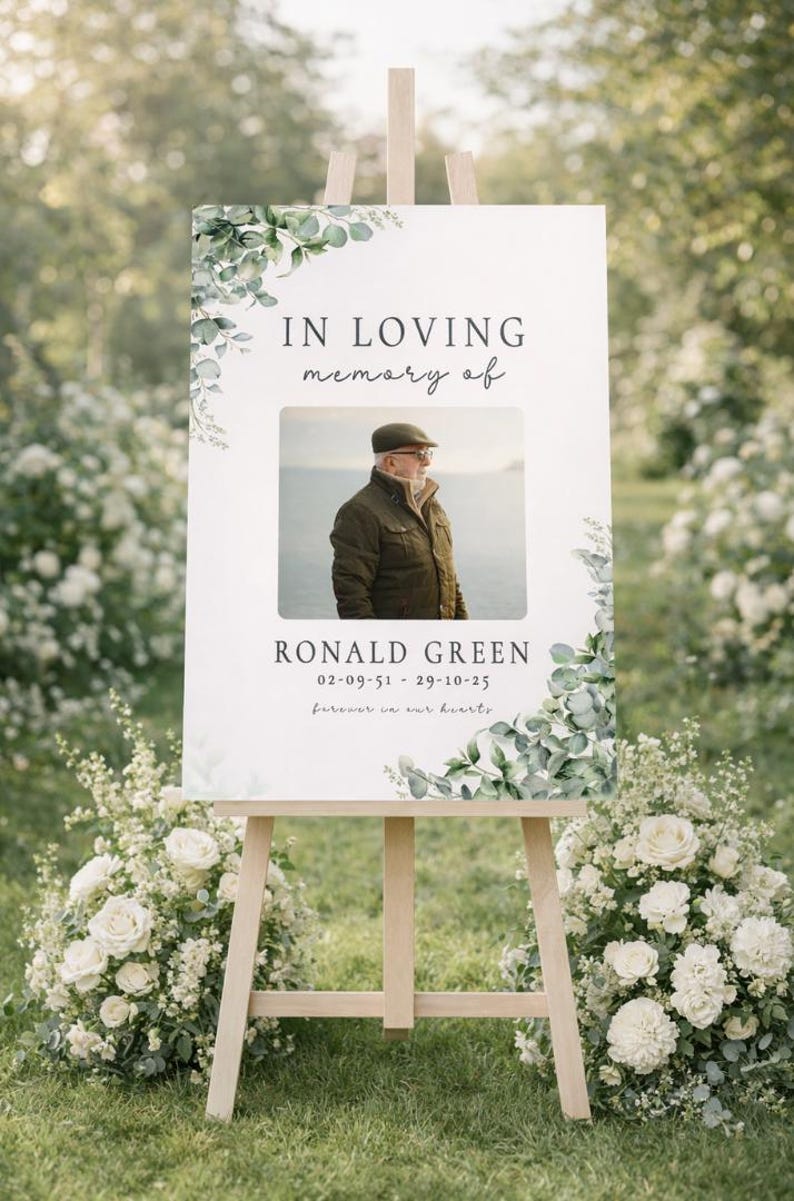 Peut inclure: Un panneau comm&eacute;moratif sur un chevalet en bois, avec le texte "IN LOVING memory of" au-dessus d'une photo d'un homme. Le panneau comprend le nom "RONALD GREEN" et des dates, avec des accents floraux. Des fleurs blanches et de la verdure entourent le panneau.