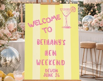May include: A yellow sign with vertical stripes, displayed on a wooden easel. The sign reads "WELCOME TO BETHANY'S HEN WEEKEND DEVON JUNE 26" in pink lettering, with a cocktail illustration. The scene suggests a party or event.