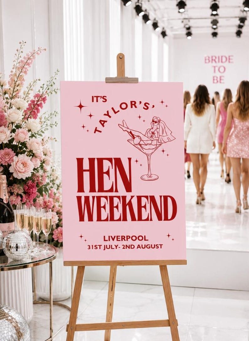 May include: A pink sign on a wooden easel reads "It's Taylor's, Hen Weekend" with a cocktail illustration and the location and dates: Liverpool, 31st July - 2nd August. In the background, women in dresses walk.