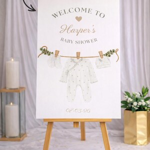 May include: A baby shower welcome sign on a wooden easel. The sign features the text "Welcome to Harper's Baby Shower" with a clothesline design holding baby clothes. The date 08.03.20 is printed at the bottom.