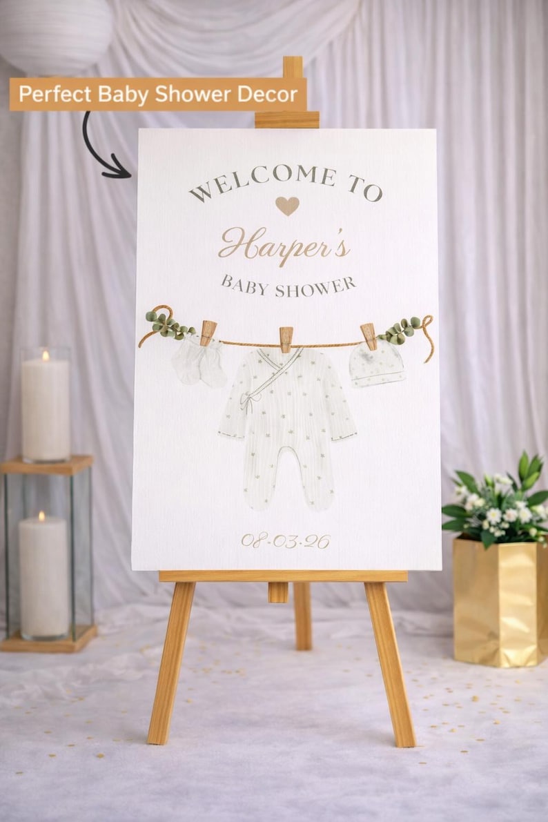 May include: A baby shower welcome sign on a wooden easel. The sign features the text "Welcome to Harper's Baby Shower" with a clothesline design holding baby clothes. The date 08.03.20 is printed at the bottom.