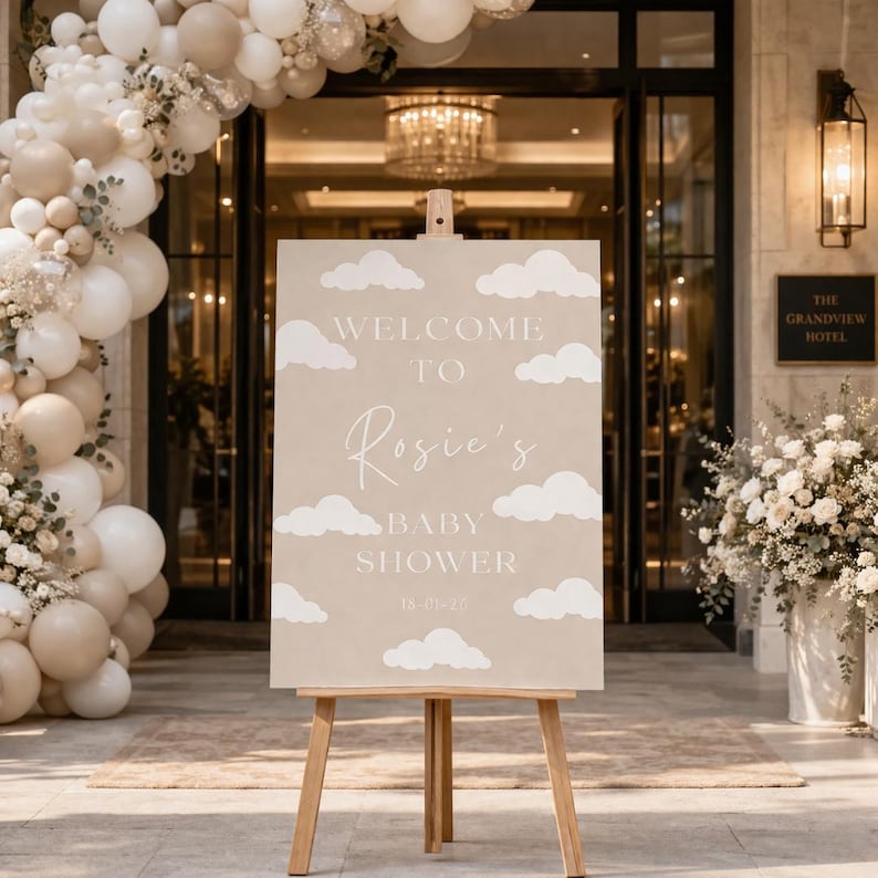 Op de afbeelding: Een beige bord op een houten ezel met de tekst "WELCOME TO Rosie's BABY SHOWER" in witte letters en wolkenillustraties. Het bord is versierd met ballonnen en bloemen, wat duidt op een babyshower-evenement.