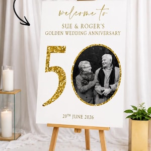 May include: A white custom anniversary board on a wooden easel. The board features gold text that reads "welcome to Sue & Roger's Golden Wedding Anniversary" and "50". A black and white photo is in the center.