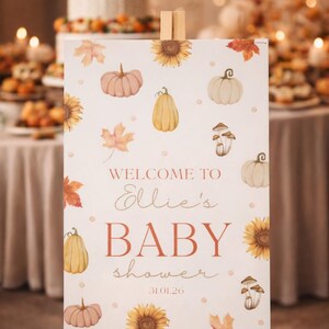 May include: A wooden easel displays a baby shower welcome sign. The sign features watercolor illustrations of pumpkins, sunflowers, and leaves. Text reads "Welcome to Ellie's Baby Shower" with the date 31.01.26.