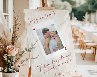 May include: A rectangular wedding sign on a wooden easel featuring a cream background with a large playing card graphic. The card displays a photo of a couple, the text Lucky in love, To love, laughter and happily ever after, and Bethany & Daniel.