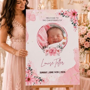 May include: A pink and white christening sign on an easel. The sign features a baby's photo surrounded by flowers, with the text "Welcome to the Christening of Louise Teller, Sunday | June 14th | 2026." The background includes floral arrangements.
