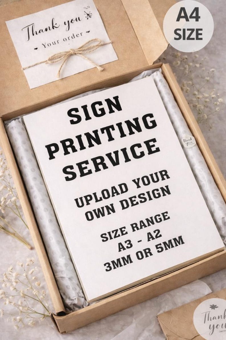 May include: A cardboard box containing a printed sign service. The sign reads "SIGN PRINTING SERVICE, UPLOAD YOUR OWN DESIGN, SIZE RANGE A3 - A2, 3MM OR 5MM." Includes a small card that says "Thank you".