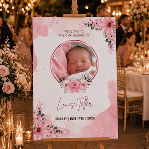 May include: A pink and white christening welcome sign on a wooden easel. The sign displays a baby's photo, floral decorations, the name "Louise Teller", and the date "14th June, 2026". The event is outdoors with fairy lights.