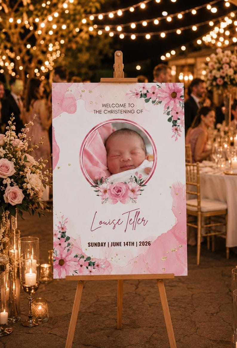 May include: A pink and white christening welcome sign on a wooden easel. The sign displays a baby's photo, floral decorations, the name "Louise Teller", and the date "14th June, 2026". The event is outdoors with fairy lights.