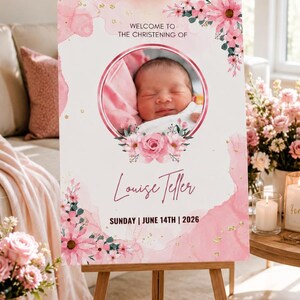 May include: A pink and white christening welcome sign on a wooden easel. It features a baby's photo, floral decorations, and the text "Welcome to the Christening of Louise Teller, Sunday | June 14th | 2026."