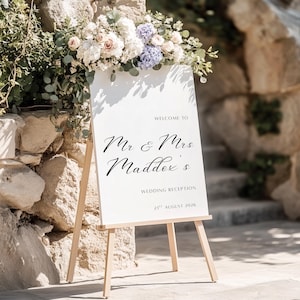 May include: A white wedding reception sign on a wooden easel, decorated with flowers. The sign says "Welcome to Mr & Mrs Maddox's Wedding Reception 21st August 2026" in black script. The sign is placed in front of a stone wall.
