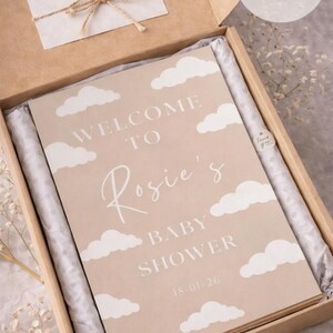 Op de afbeelding: A4-formaat babyborrelbord in een kartonnen doos. Het bord is beige met witte wolkenillustraties en de tekst "WELCOME TO Rosie's BABY SHOWER 18-01-26". Een kleine kaart met een strik en de woorden "Thank you" is ook inbegrepen.