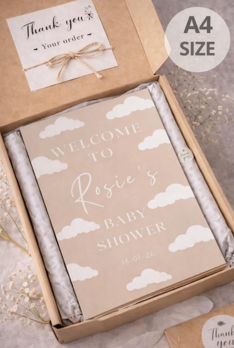 Op de afbeelding: A4-formaat babyborrelbord in een kartonnen doos. Het bord is beige met witte wolkenillustraties en de tekst "WELCOME TO Rosie's BABY SHOWER 18-01-26". Een kleine kaart met een strik en de woorden "Thank you" is ook inbegrepen.