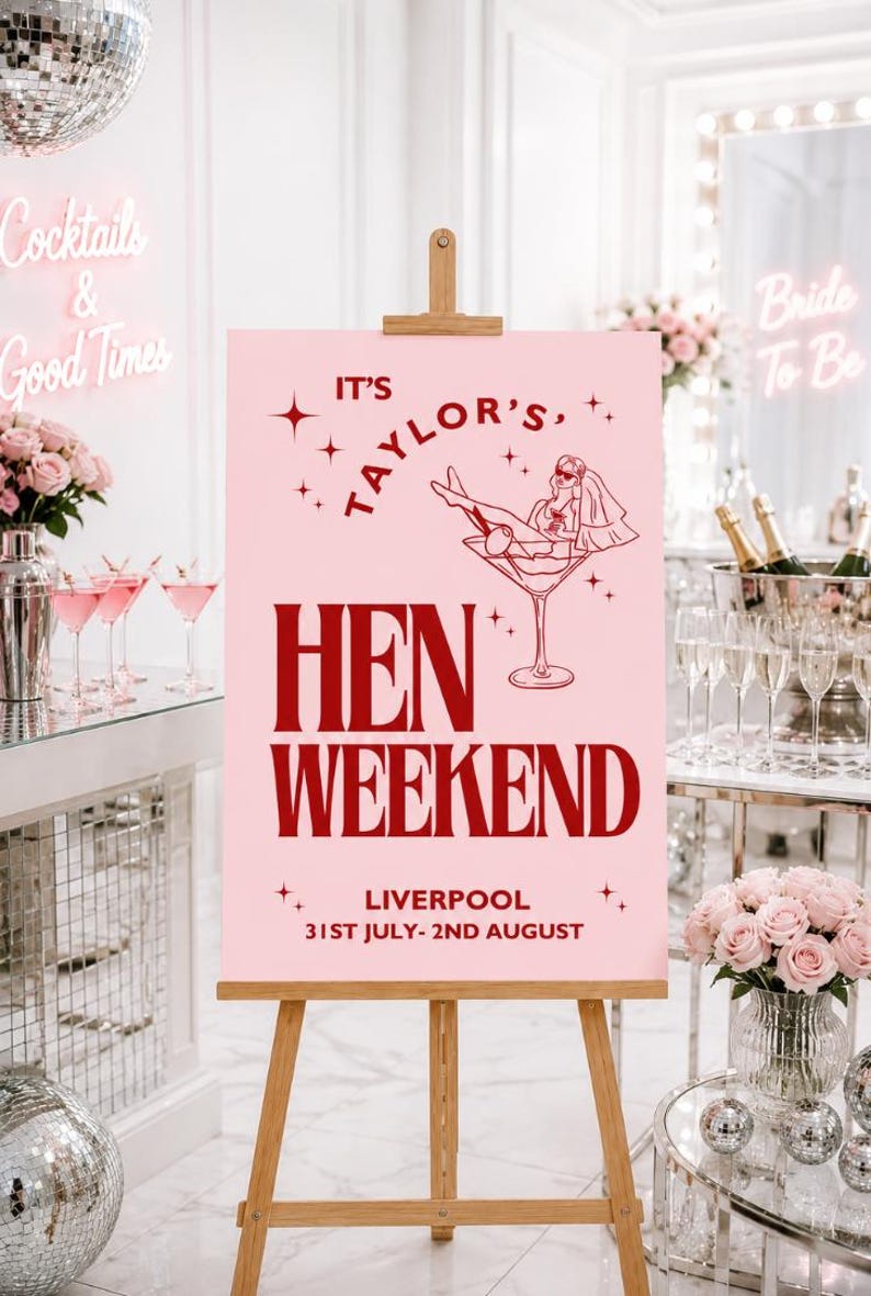 May include: A pink sign on a wooden easel with the text "It's Taylor's Hen Weekend Liverpool 31st July-2nd August". The sign features a graphic of a woman in a martini glass. The background includes cocktails and flowers.