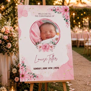 May include: A pink and white christening sign on a wooden easel. The sign displays "Welcome to the Christening of Louise Teller" and the date "Sunday | June 14th | 2026." A baby's photo is in the center, framed by floral decorations.