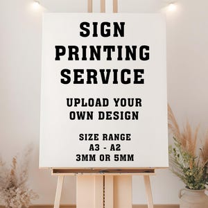 May include: A white sign on an easel with the text "SIGN PRINTING SERVICE". The sign also reads "UPLOAD YOUR OWN DESIGN" and "SIZE RANGE A3 - A2 3MM OR 5MM". The easel is made of wood and is in a well-lit room.
