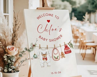 May include: A white welcome sign for Chloe's baby shower on a wooden easel. The sign features red text, a small red heart, and a clothesline illustration with a baby onesie, a bib, a gingerbread man, and a red knit hat. The date 13-10-26 is printed.