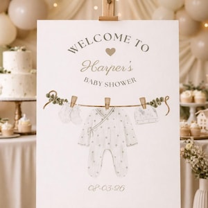 May include: A baby shower sign on an easel with the text "Welcome to Harper's Baby Shower." The sign features a watercolor illustration of baby clothes hanging on a clothesline, including a onesie, hat, and mittens. The date "08-03-26" is printed at the bottom.