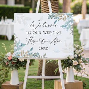 May include: A white wedding welcome sign with green and brown watercolour leaves and the text "Welcome to our wedding Rosie & Alex 23.07.25".