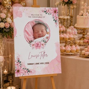 May include: A christening welcome sign with a baby's photo in a pink floral frame. The sign displays the name "Louise Teller" and the date "June 14th, 2026". The sign is on a wooden easel, with pink and gold balloons and floral arrangements in the background.