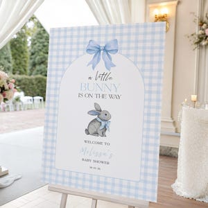 May include: A rectangular welcome sign featuring a light blue and white gingham pattern. The centre has an arched white panel with a blue bow, a grey bunny illustration, and the text: A little BUNNY IS ON THE WAY WELCOME TO Melissa's BABY SHOWER 10-03-26.