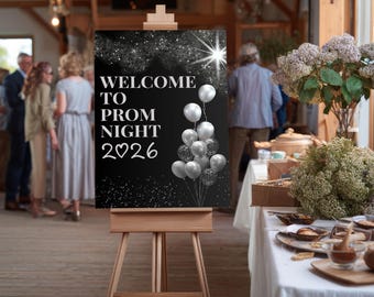 May include: A black sign on a wooden easel with the words "WELCOME TO PROM NIGHT 2026" in white. Silver balloons and starbursts decorate the sign. The background shows a party setting with people and tables.