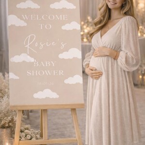 Op de afbeelding: Een beige babyborrelbord op een ezel met witte wolken en de tekst "Welcome to Rosie's Baby Shower". Een vrouw in een cr&egrave;mekleurige jurk staat naast het bord. Datum: 18-01-26.