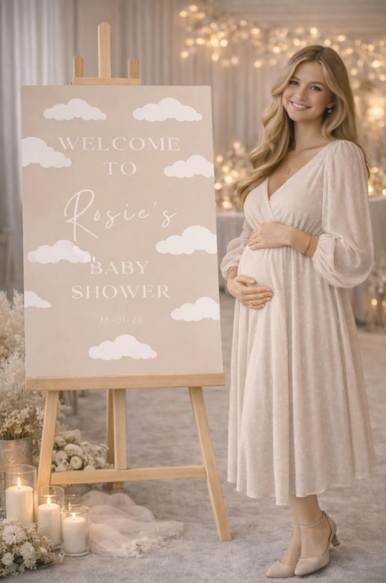 Op de afbeelding: Een beige babyborrelbord op een ezel met witte wolken en de tekst "Welcome to Rosie's Baby Shower". Een vrouw in een cr&egrave;mekleurige jurk staat naast het bord. Datum: 18-01-26.