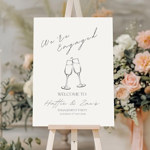 May include: A rectangular white engagement party sign on a wooden easel featuring black text that reads We're Engaged, Welcome to Hattie & Zac's Engagement Party, and Saturday 2nd May 2026, with a black line drawing of two clinking champagne flutes.