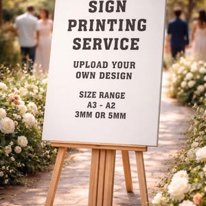 May include: A wooden easel displays a white sign with black text: "SIGN PRINTING SERVICE, UPLOAD YOUR OWN DESIGN, SIZE RANGE A3 - A2, 3MM OR 5MM." The sign is set on a stone path lined with white roses, with blurred figures in the background.