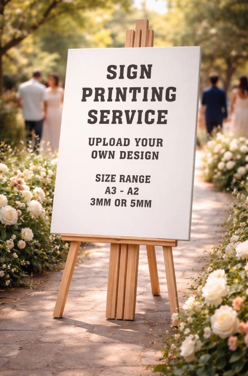May include: A wooden easel displays a white sign with black text: "SIGN PRINTING SERVICE, UPLOAD YOUR OWN DESIGN, SIZE RANGE A3 - A2, 3MM OR 5MM." The sign is set on a stone path lined with white roses, with blurred figures in the background.